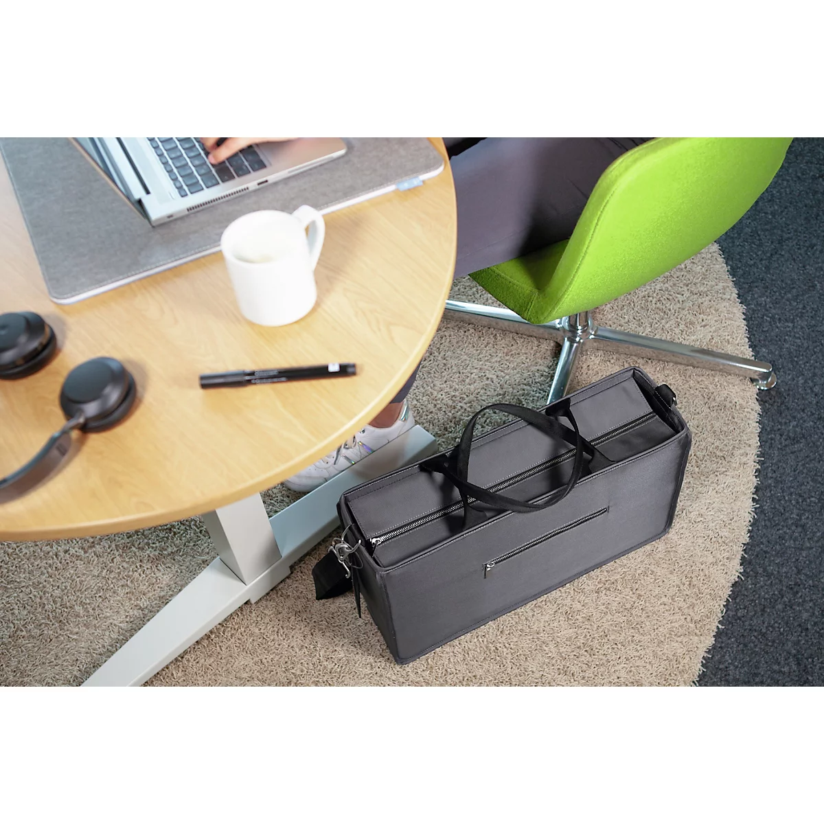 Sac noir au sol, table avec ordinateur portable, tasse à café, écouteurs. Personne assise sur une chaise verte.