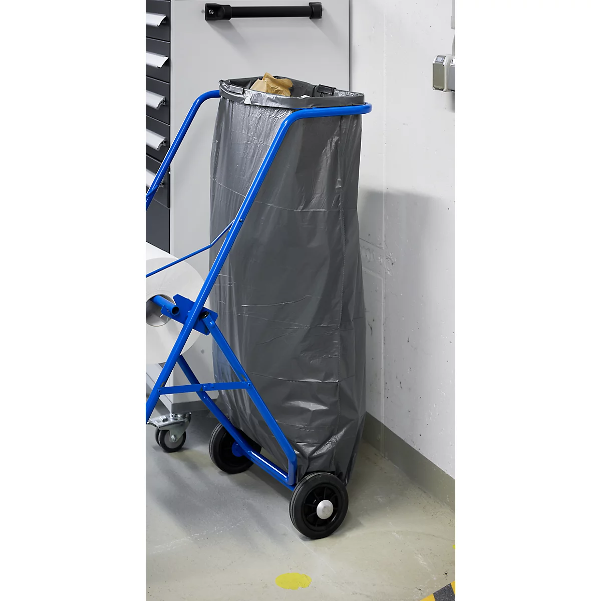 Sac poubelle gris dans un chariot bleu. Le chariot se trouve dans une pièce avec un fond blanc.