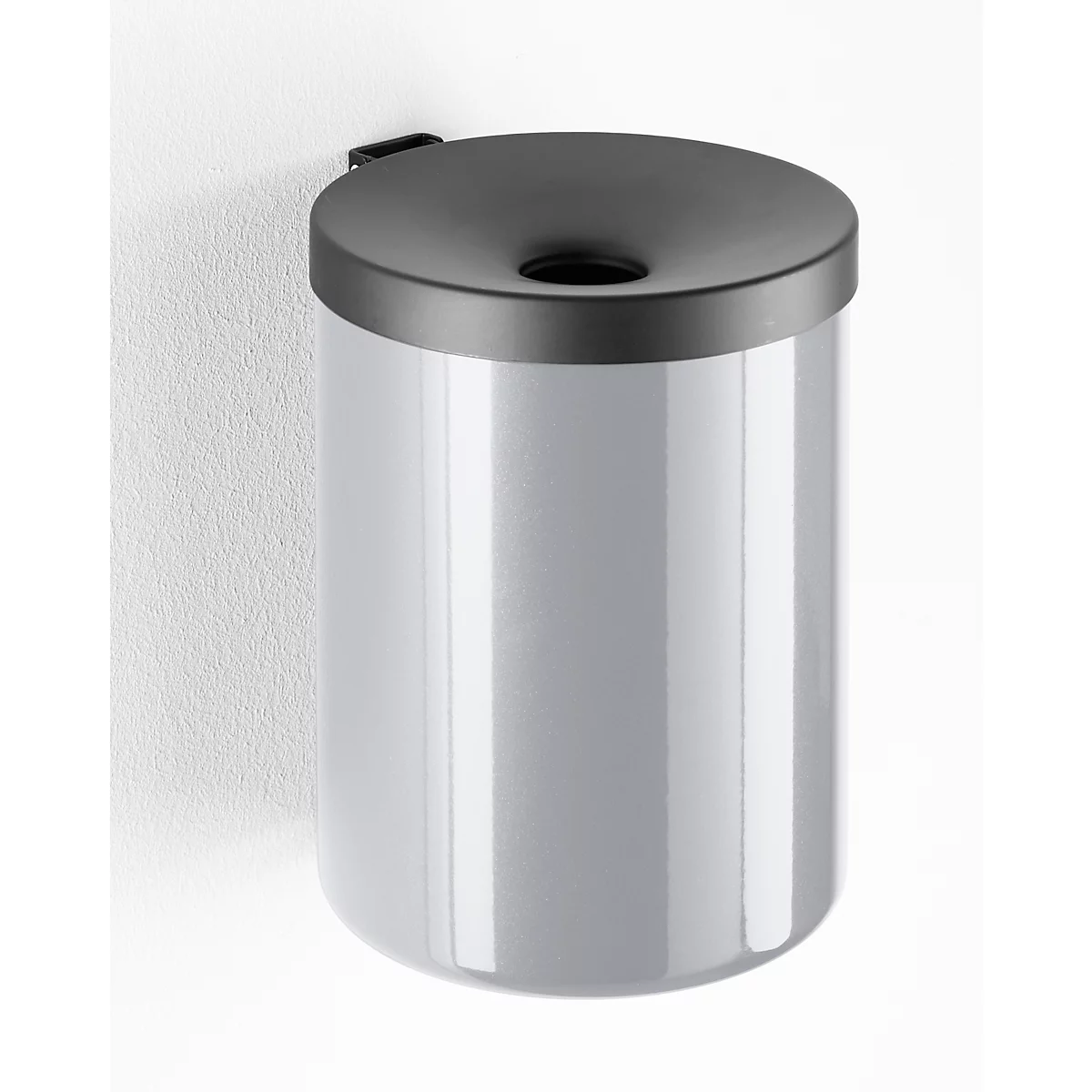 Gray trash can on white wall. The lid is black with a round opening. The bin is cylindrical.