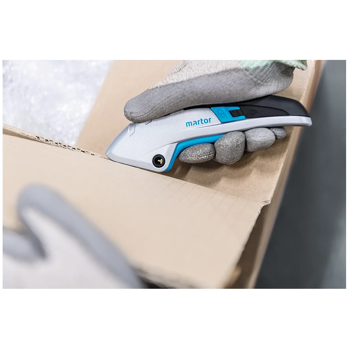 A gloved hand cutting open a cardboard box with a cutter. The cutter has a blue accent and the writing martor.