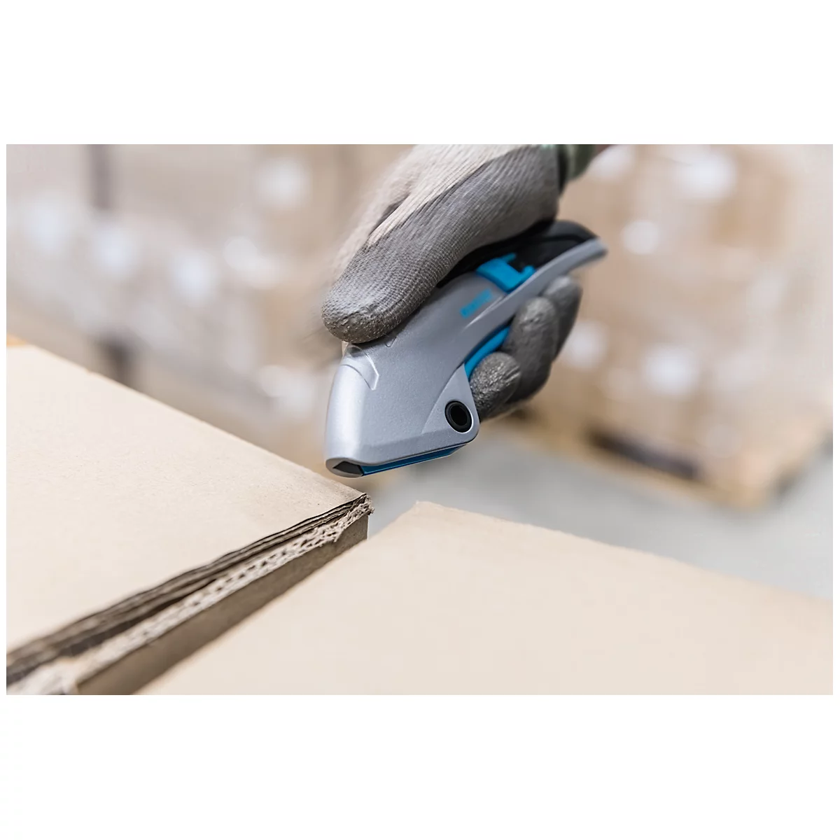 A worker cuts cardboard with a gray and blue cutter. The cardboard is open. His hand wears a gray glove.