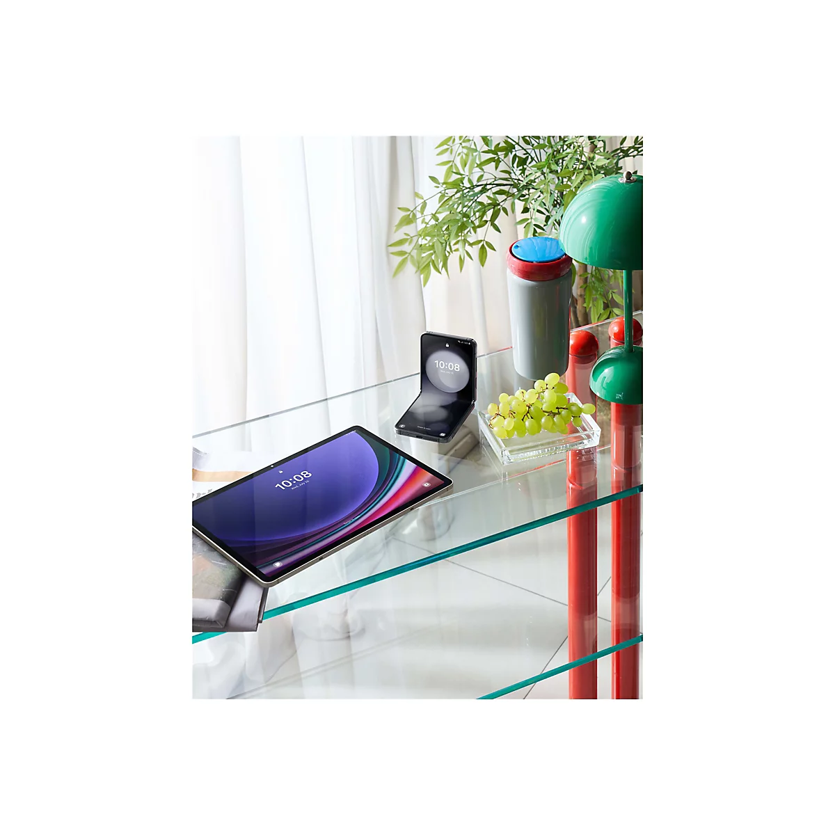 Ein Tablet und ein Smartphone liegen auf einem Glastisch. Eine Traube und eine Lampe stehen daneben. Der Hintergrund ist hell.