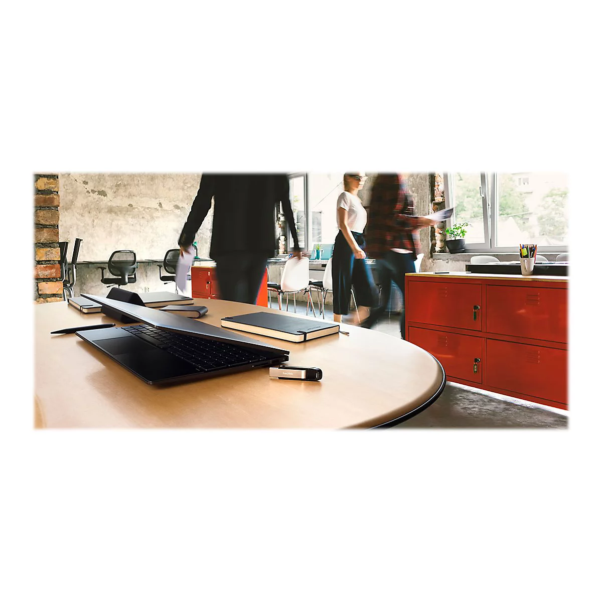 Un ordinateur portable noir ouvert sur une table ronde en bois. En arrière-plan, des personnages flous marchent dans un bureau.