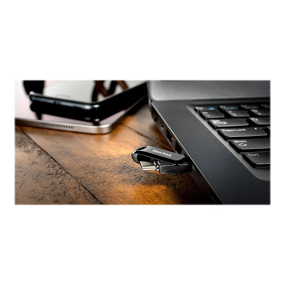 Ein schwarzer SanDisk USB-Stick steckt in einem Laptop, daneben ein Smartphone, beides auf Holz.