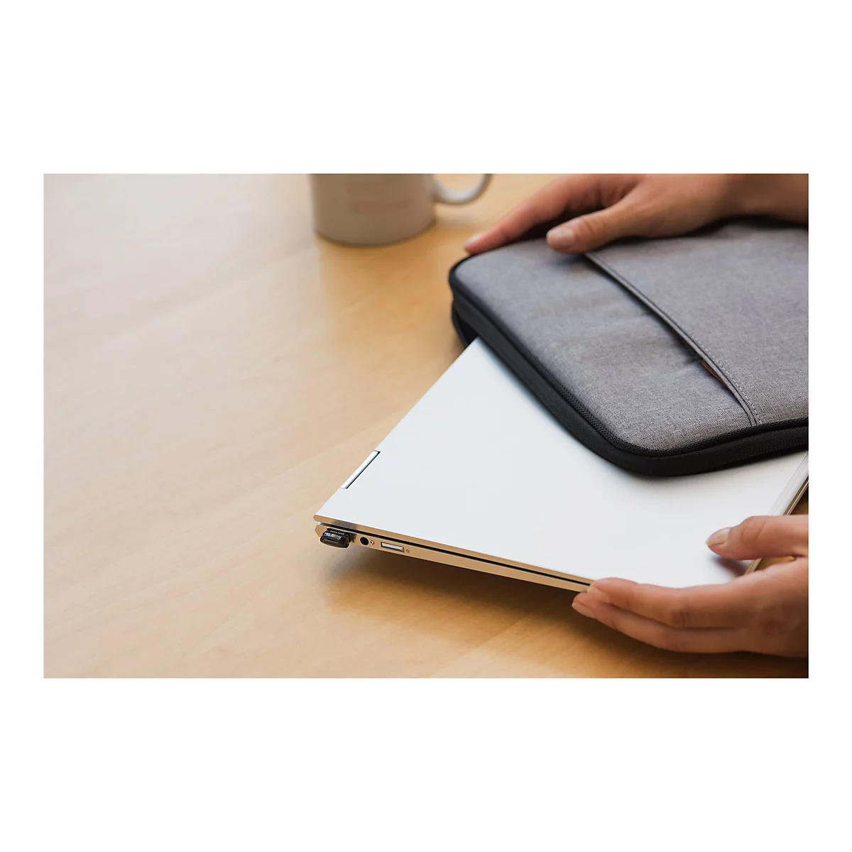 Des mains tiennent une housse d'ordinateur portable grise sur un ordinateur portable argenté. Une tasse est posée sur la table en bois.