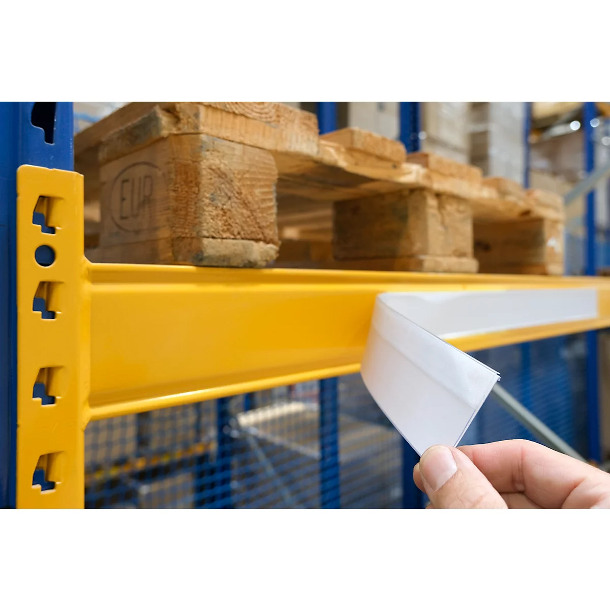 Hand houdt een wit etiket op een gele legplank. Boven, pallets. Magazijn op de achtergrond.