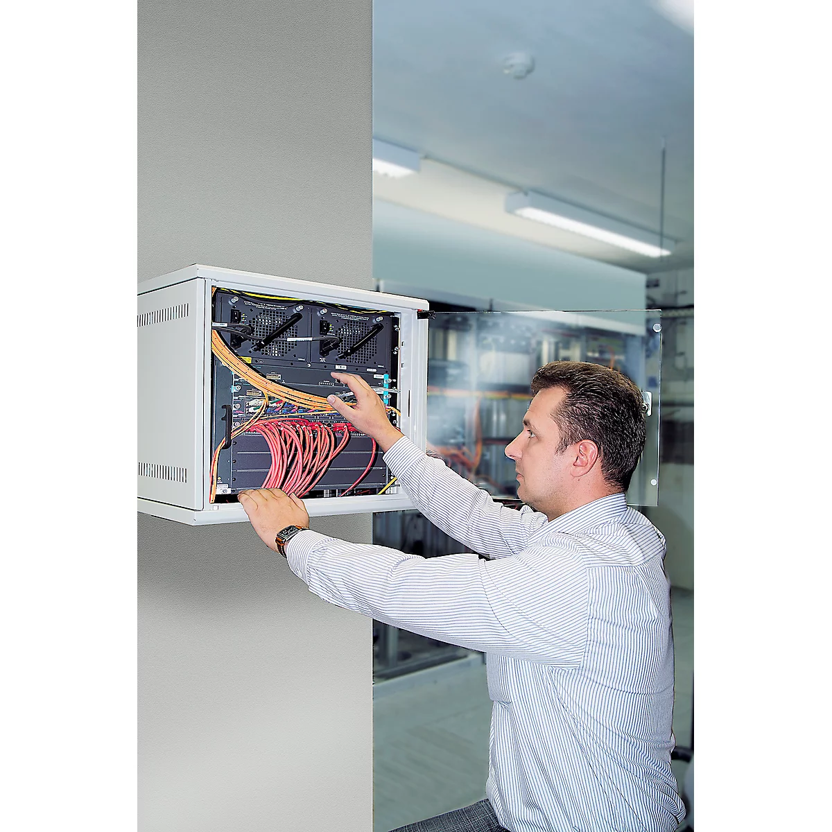 A man works on a server cabinet. He connects red and yellow cables. The cabinet is mounted in a wall.