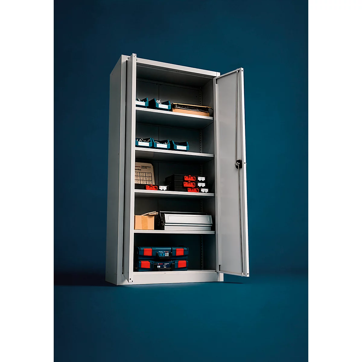 Une armoire métallique grise ouverte sur fond bleu foncé, remplie de mallettes à outils et de boîtes de rangement.