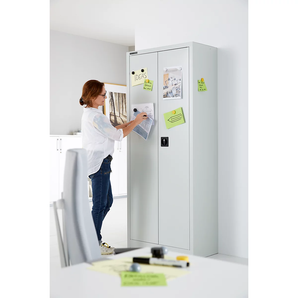 Une femme en jean et chemisier blanc fixe des notes sur une armoire blanche. Sur les notes, on peut lire "idées".