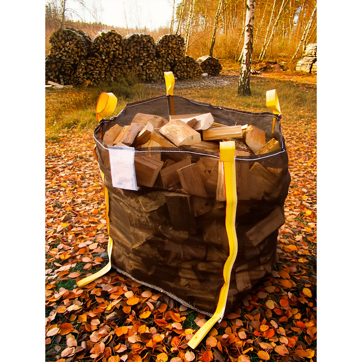 Un grand sac rempli de bois est posé sur des feuilles. Des sangles de transport jaunes sont visibles. En arrière-plan, des tas de bois.