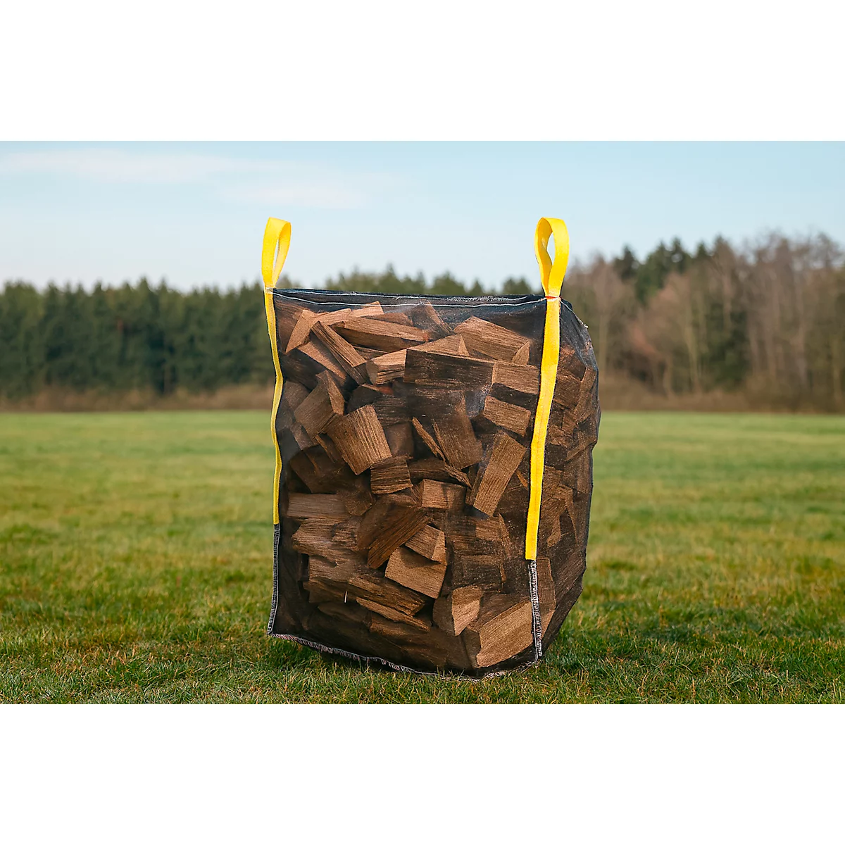 Un grand sac noir rempli de bois de chauffage se trouve sur une prairie verte. Des boucles jaunes servent de poignées.