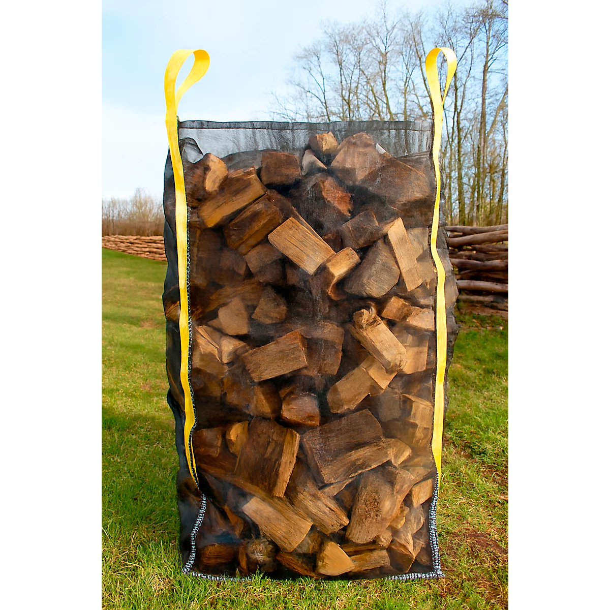 Un grand sac rempli de bois, avec des poignées de transport jaunes, se dresse à l'extérieur sur une prairie.