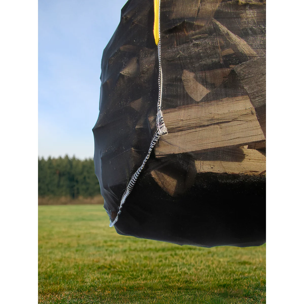 Zwarte zak vol houtblokken. Gele riem om vast te zetten, gestikt met witte draad. Op de achtergrond weide en bos.