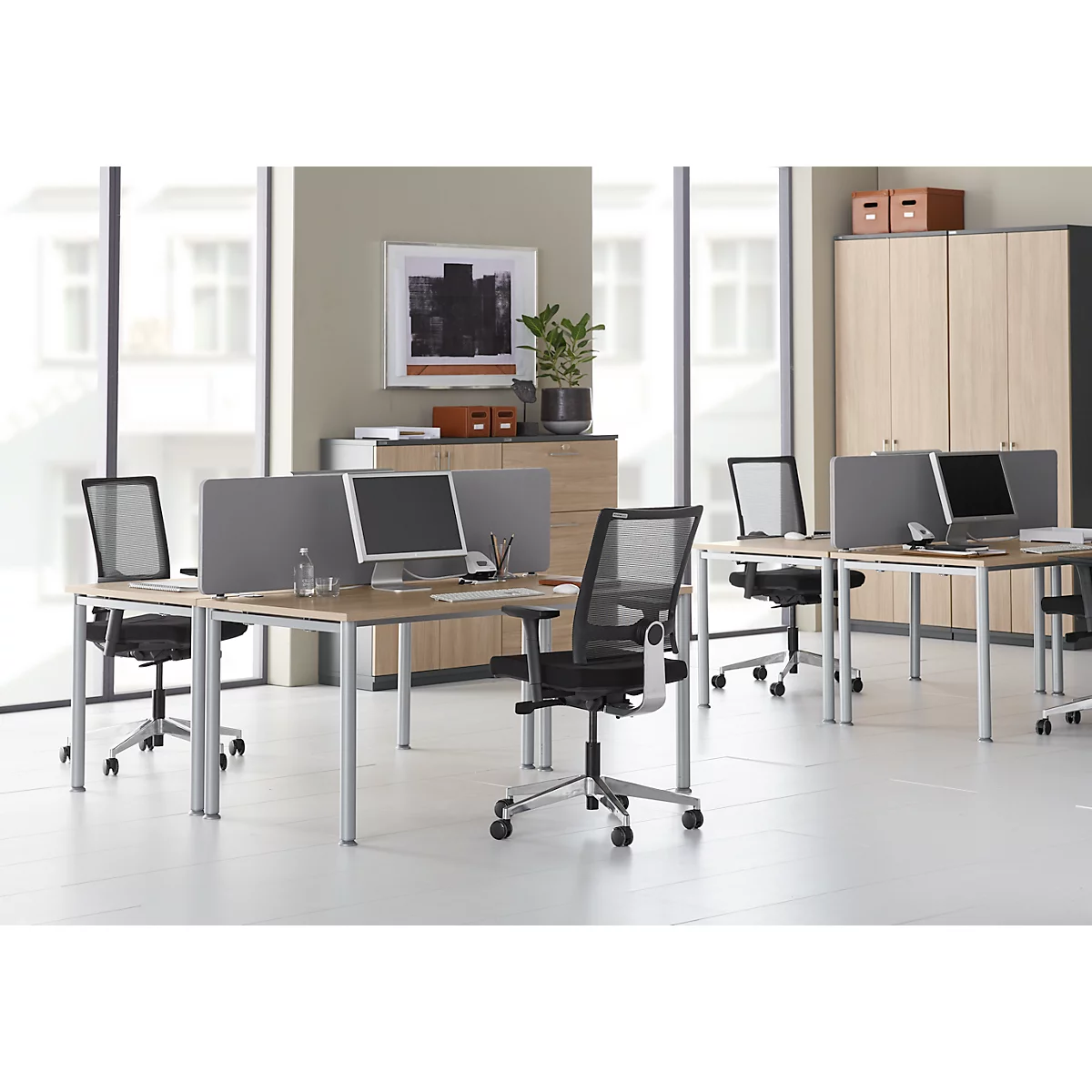 Kantoor met meerdere bureaus, stoelen en computers. Lichte omgeving met kasten en grote ramen.