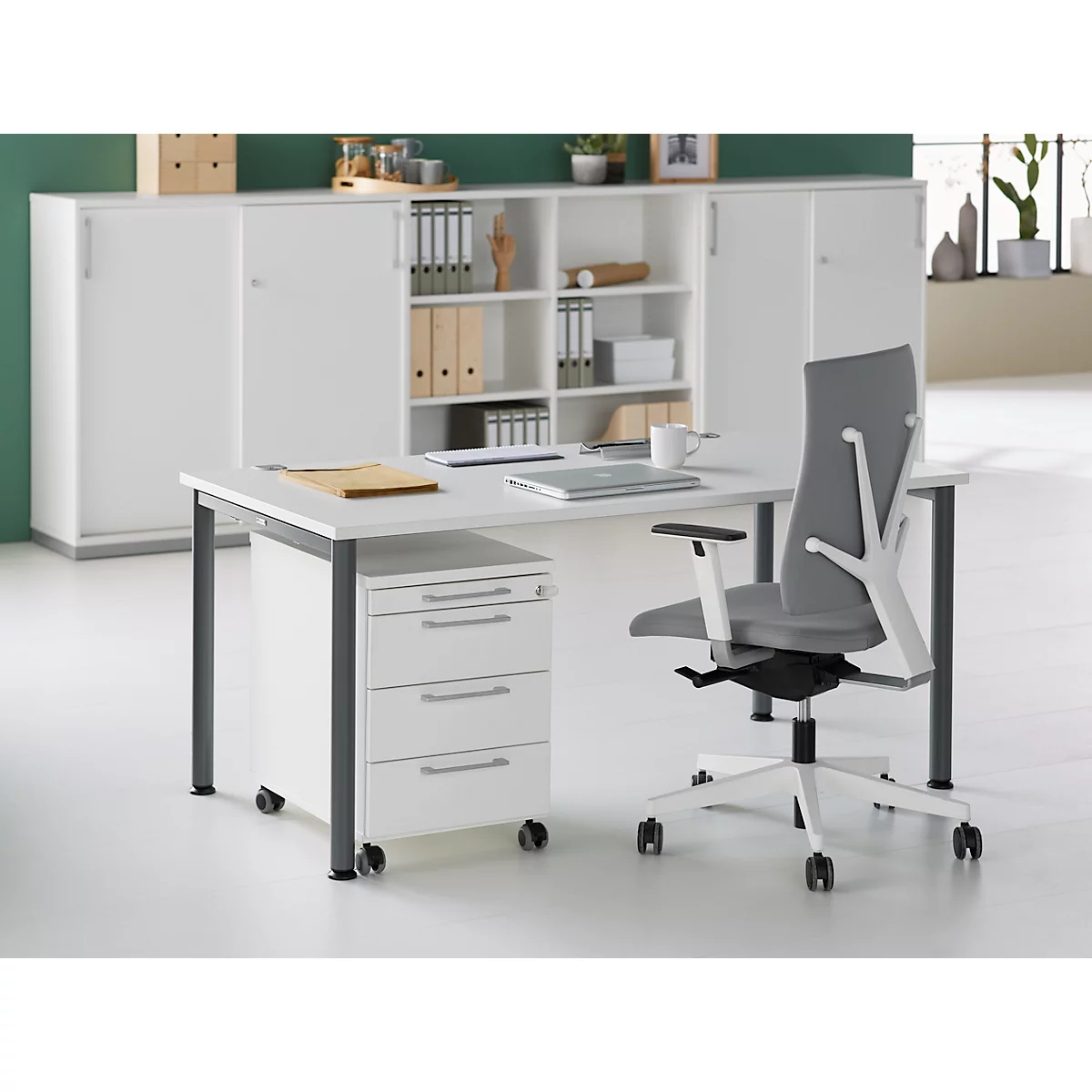 Een modern bureau met een grijze bureaustoel. Achter het bureau staat een witte plank en kast.