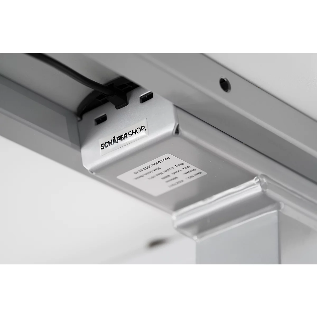 Close-up van de onderkant van een tafel. Op een metalen plaat staat 'SCHÄFER SHOP'. Kabels en een sticker zijn zichtbaar.