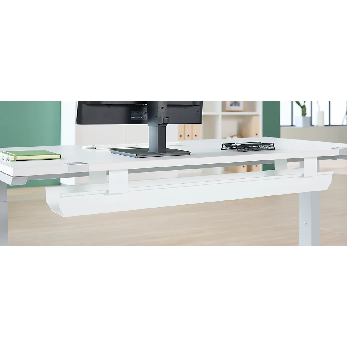 Bureau blanc avec passe-câbles intégré et moniteur. Un carnet vert et un porte-stylos sont sur le dessus de la table.