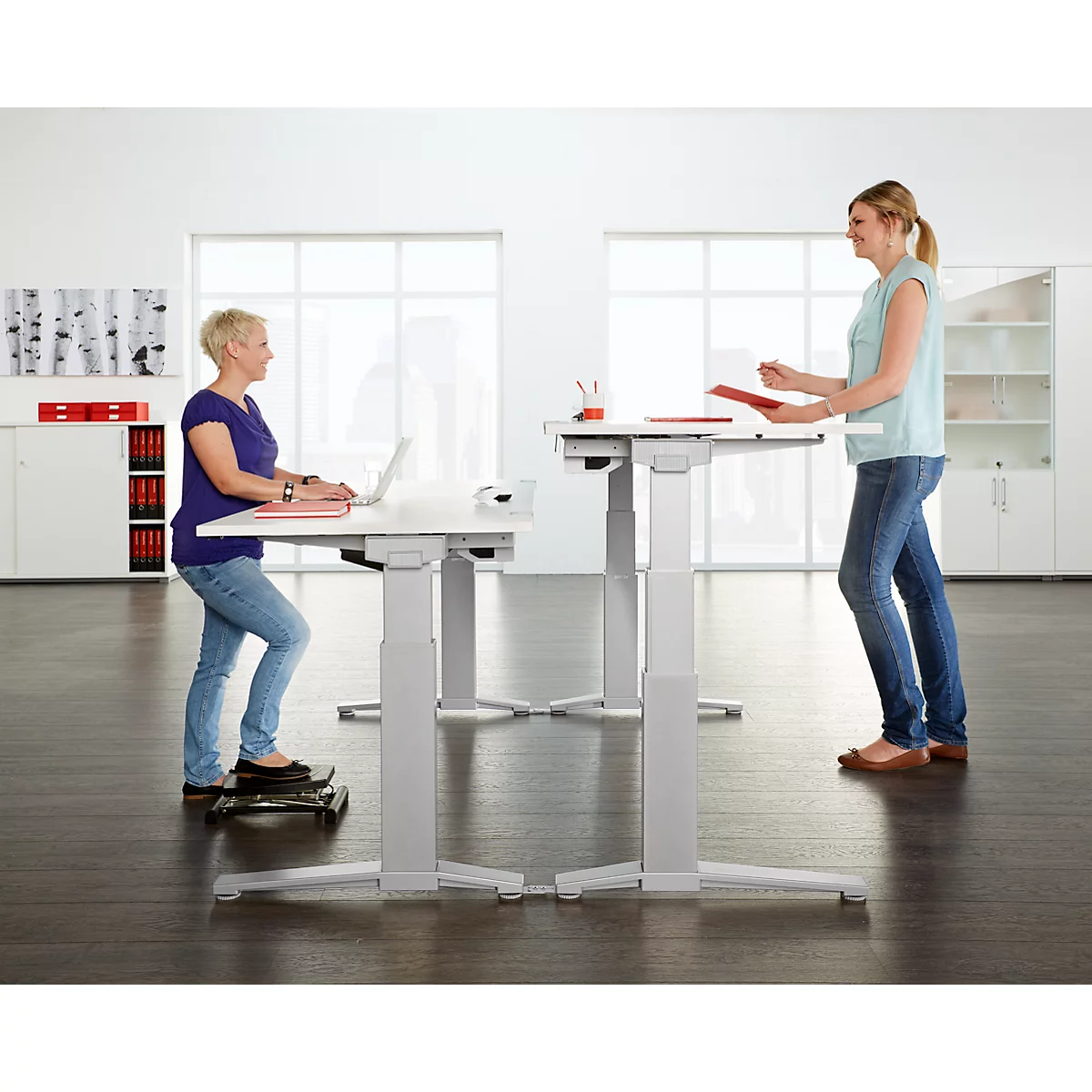 Two women standing and working at height-adjustable desks in a bright office.