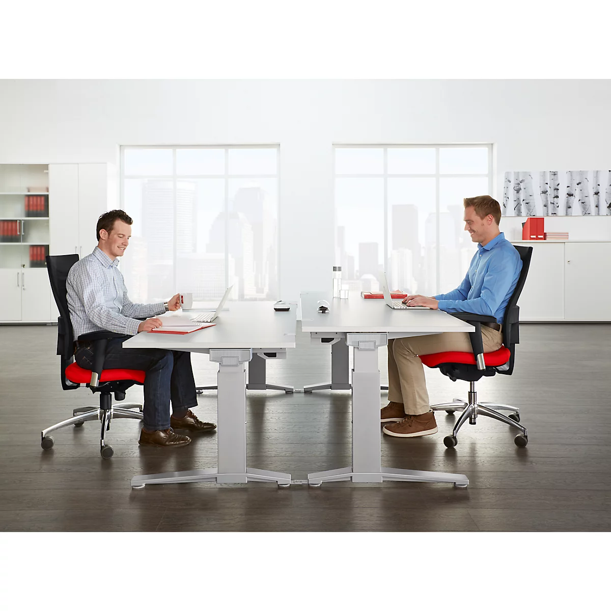 Two men sitting at height-adjustable desks, working on laptops, smiling.
