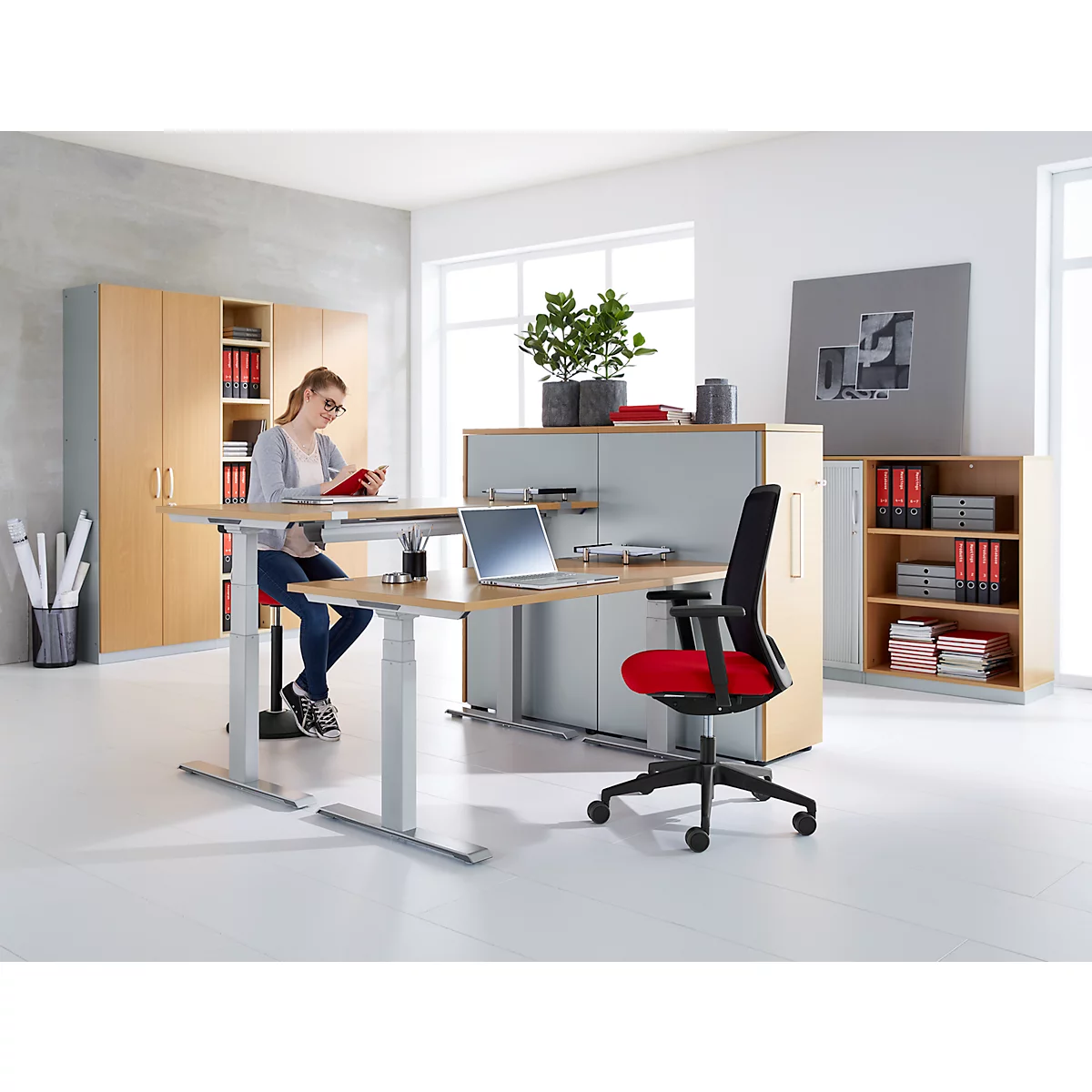 Woman seated at height-adjustable desk, reading, laptop. Office chair with red seat. Brightly furnished.