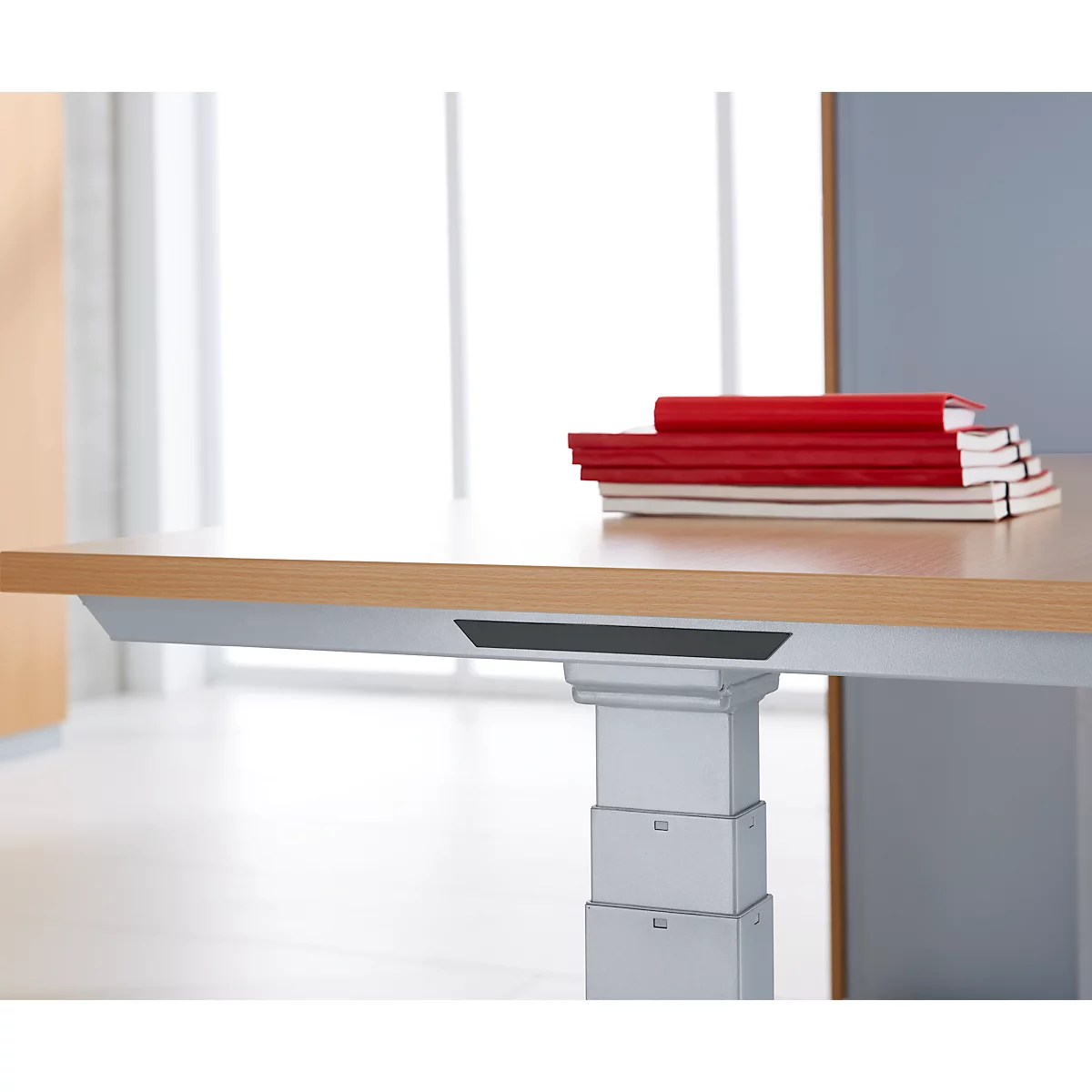 A desk with a wooden top and a silver frame. On it are red and white notebooks. Windows in the background.