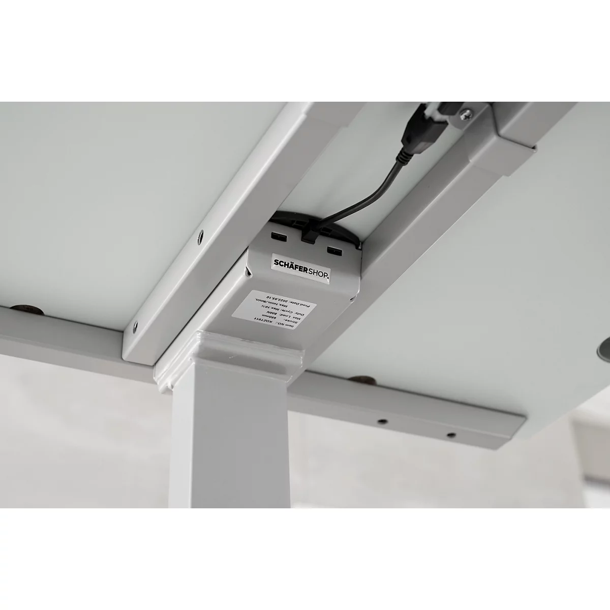 Close-up of the underside of a height-adjustable desk. A gray metal frame supports the table top with a label. The cables are connected.