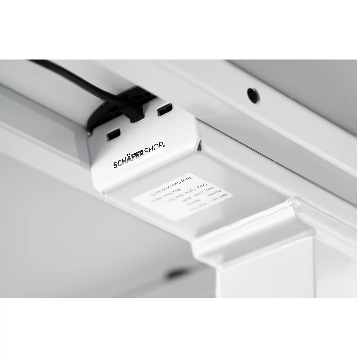 Close-up of the underside of a white desk. A black cable leads to a white connector printed with "SCHÄFER SHOP".