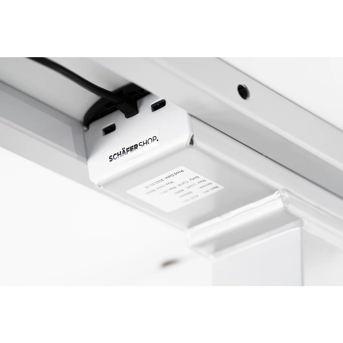 Close-up of the underside of a white desk. A white housing with the lettering "SCHÄFER SHOP" and a sticker is shown. A black cable leads into a hole in the table.