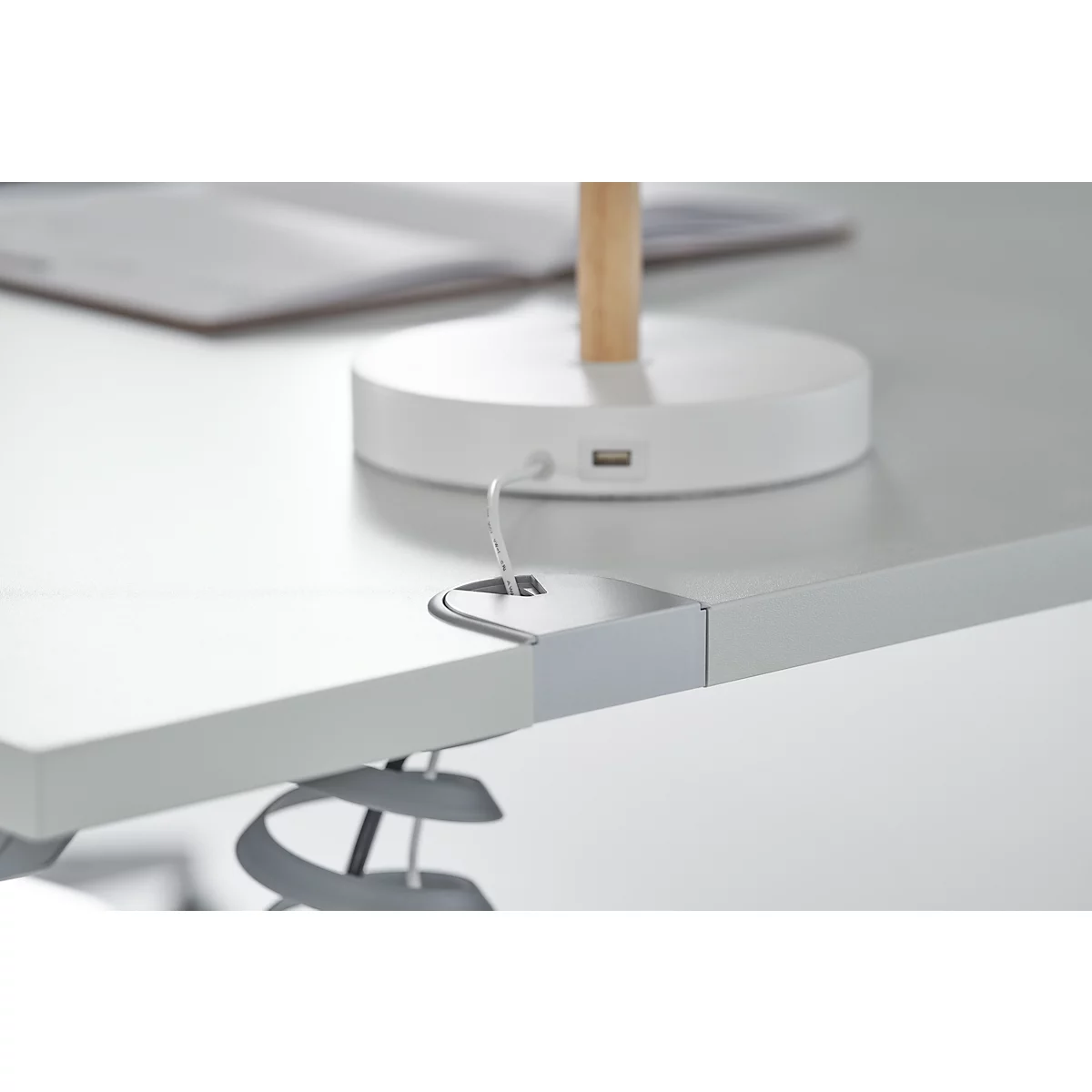 Close-up of a white desk. Part of a table lamp is visible, equipped with a USB port. A book lies on the table.