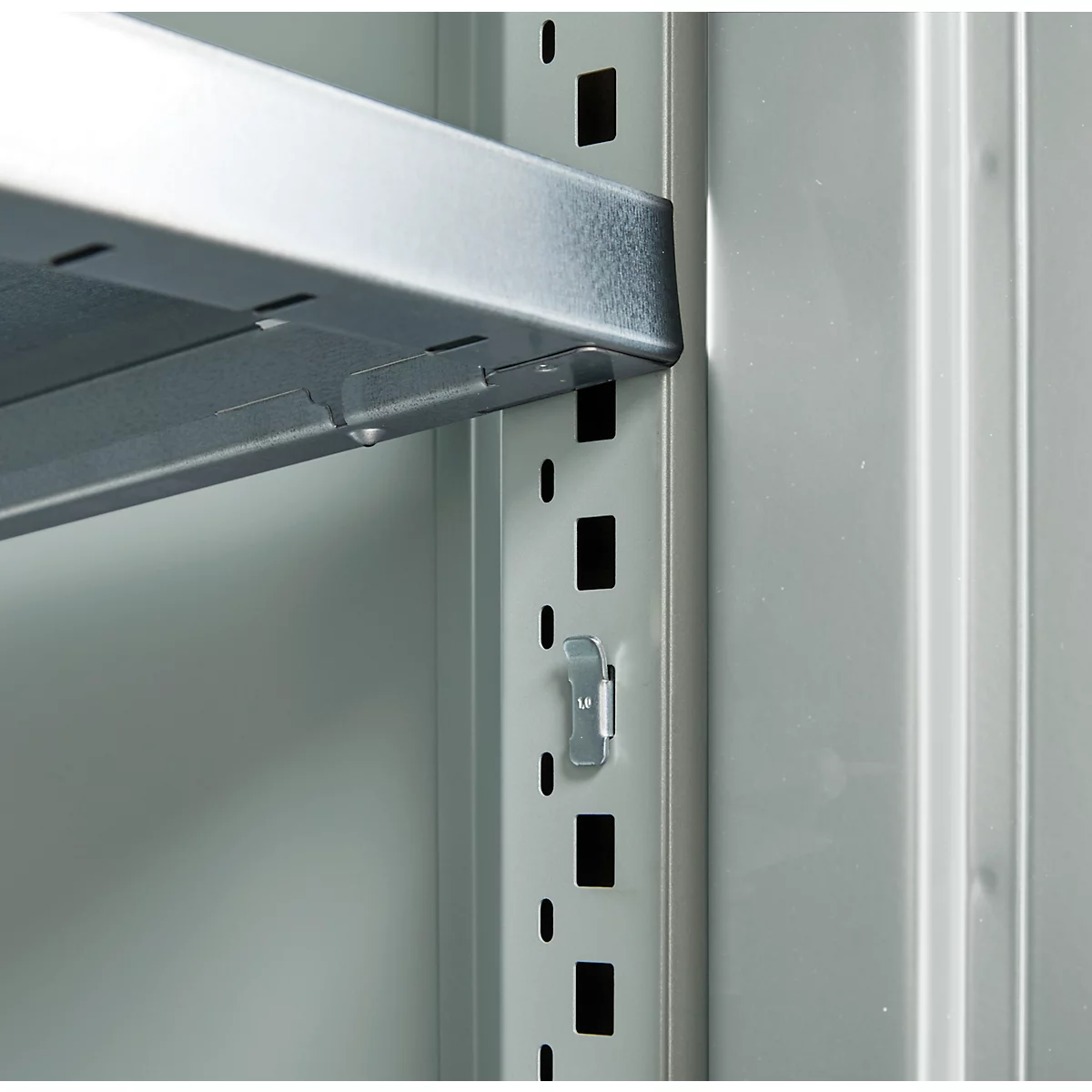 Close-up of a shelving system. A silver shelf is inserted into a holder, next to a series of holes and brackets.