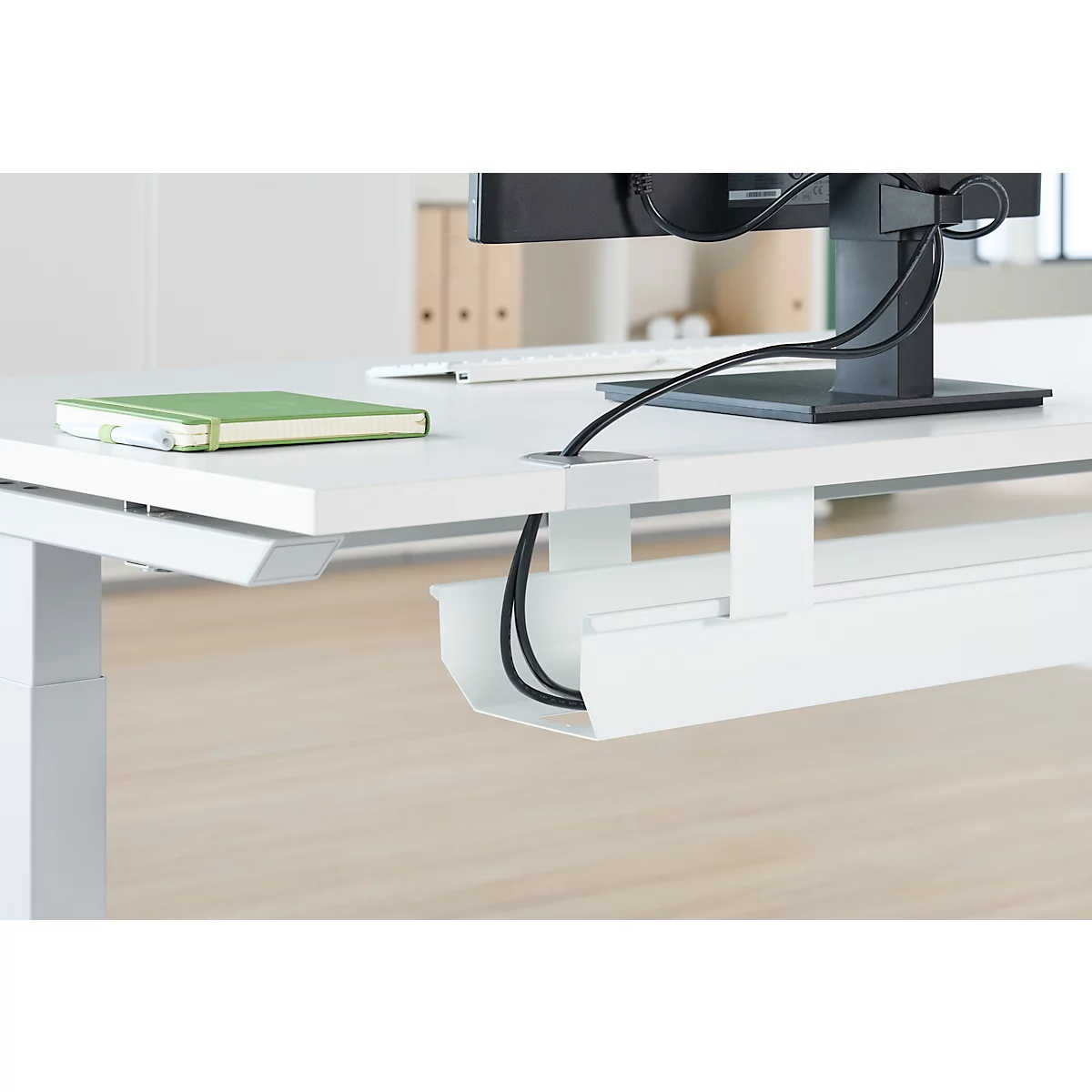 Nahaufnahme eines weissen Schreibtisches mit Kabelmanagement, Monitor und Notizbuch.