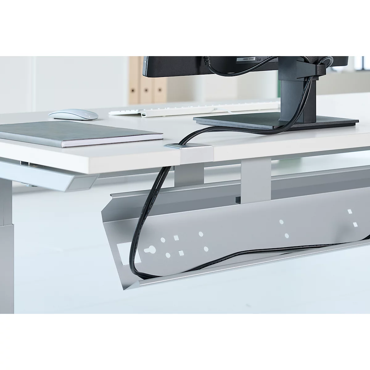 Detailansicht eines Schreibtisches mit Kabelwanne. Schwarze Kabel führen zu Monitor und Stromanschluss. Graue Kabelwanne unter der Tischplatte.