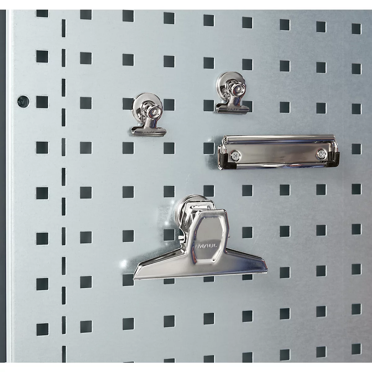 Metallklammern und eine Klemmbrettklammer an einer Lochwand. Auf dem silberfarbenen Hintergrund sind quadratische Löcher angeordnet.