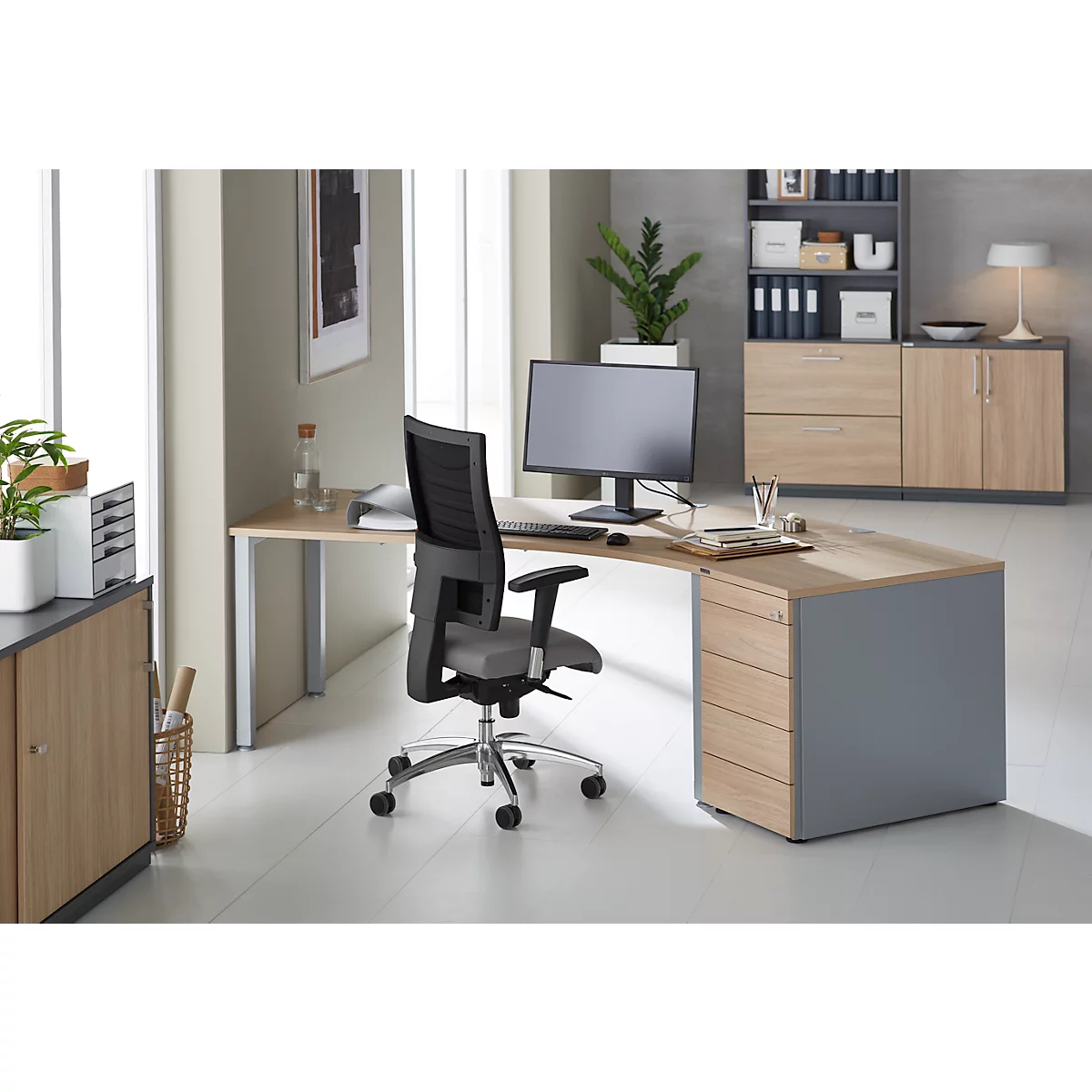 A modern corner desk with office chair in a bright office. On the desk are a monitor, plants and office supplies.