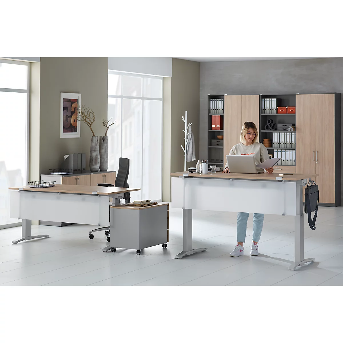 Woman at height-adjustable desk with laptop, documents. Office scene with cabinets and window.
