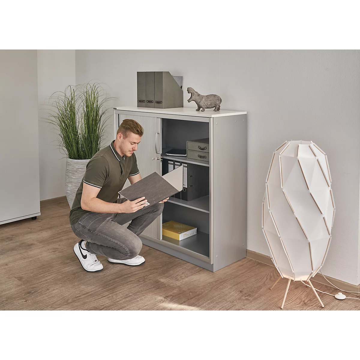 Ein Mann kniet vor einem grauen Aktenschrank, um Aktenordner zu sichten. Rechts eine moderne Stehlampe.