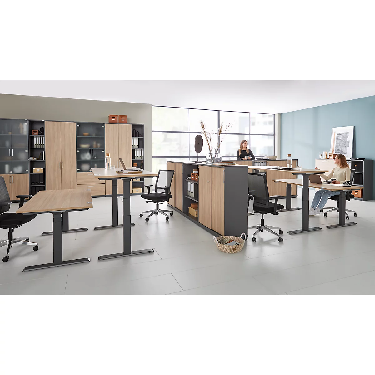Büro mit höhenverstellbaren Schreibtischen, Schränken und zwei Personen, die an Laptops arbeiten.