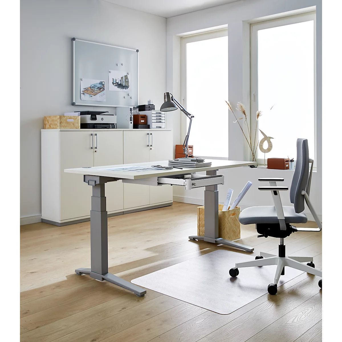 Heller Büroraum mit höhenverstellbarem Schreibtisch, Bürostuhl und Fenster. Auf dem Tisch liegen Unterlagen und eine Lampe.