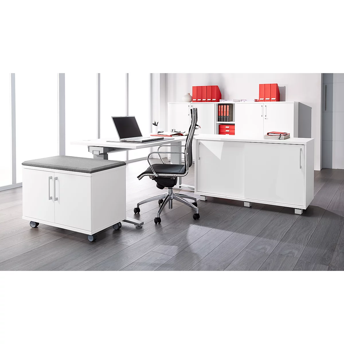 Office with white desk, chair, laptop, wall unit and roll container. Grey floor, lots of daylight.