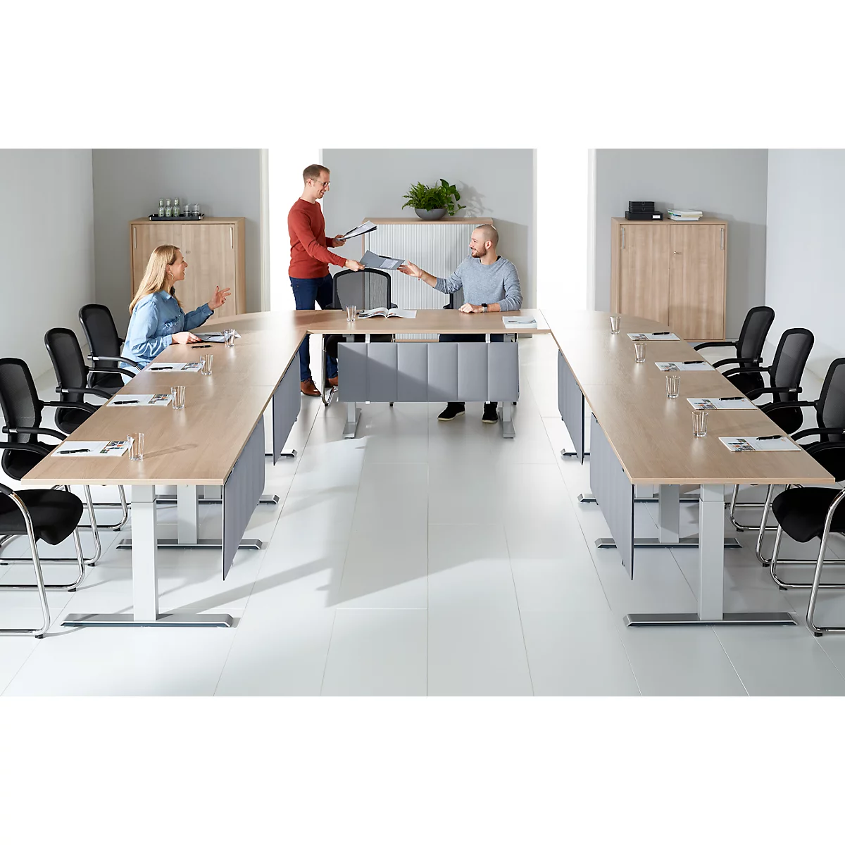 Salle de conférence avec des meubles clairs. Trois personnes sont assises à une table en forme de U, une personne remet des papiers.