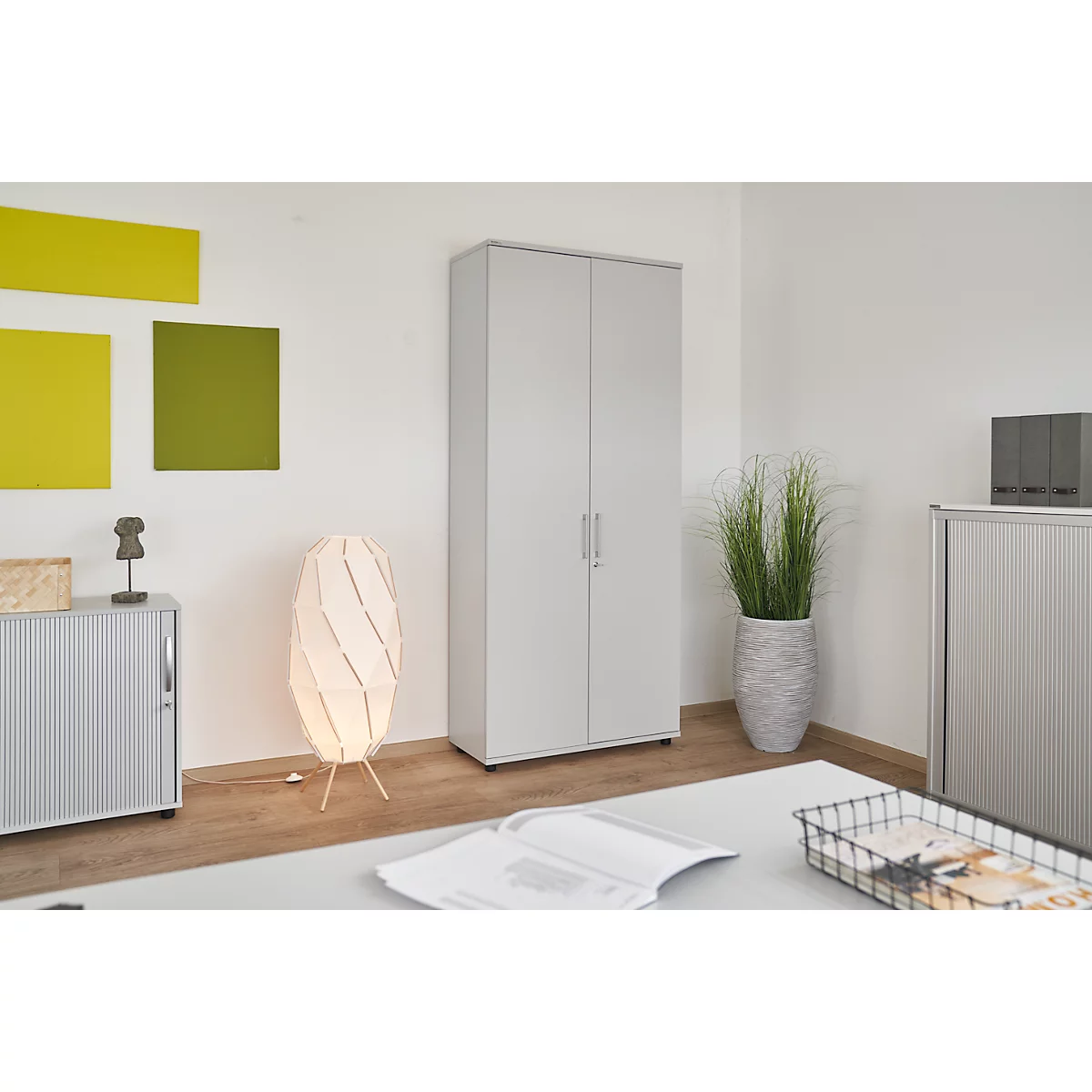 Bright cabinet in the office. Next to it a floor lamp and a large flower pot with grass. On the desk is an open book.