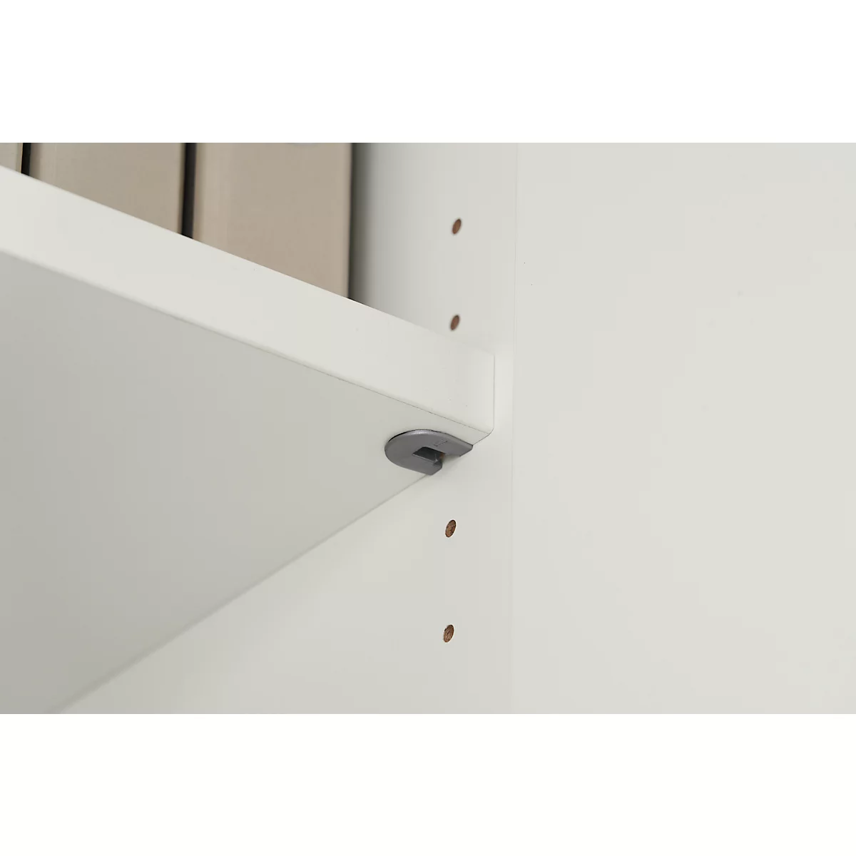 Close-up of a white shelf board with grey bracket. Below a white wall with pre-drilled holes.