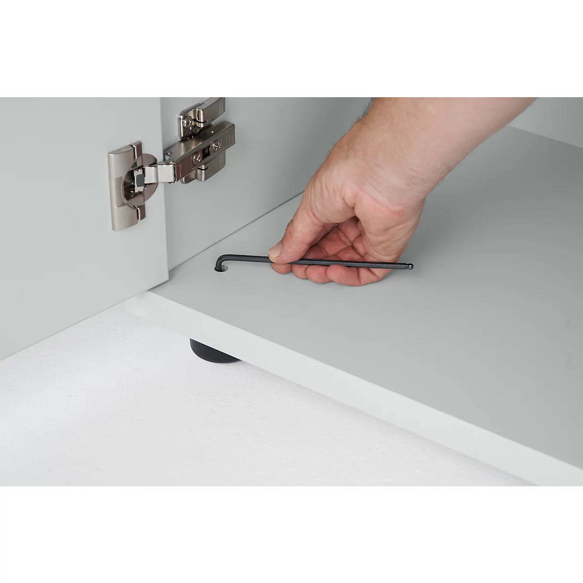 Hand holding an Allen key over a gray surface with a hinge on a cabinet door. A black bolt is visible.