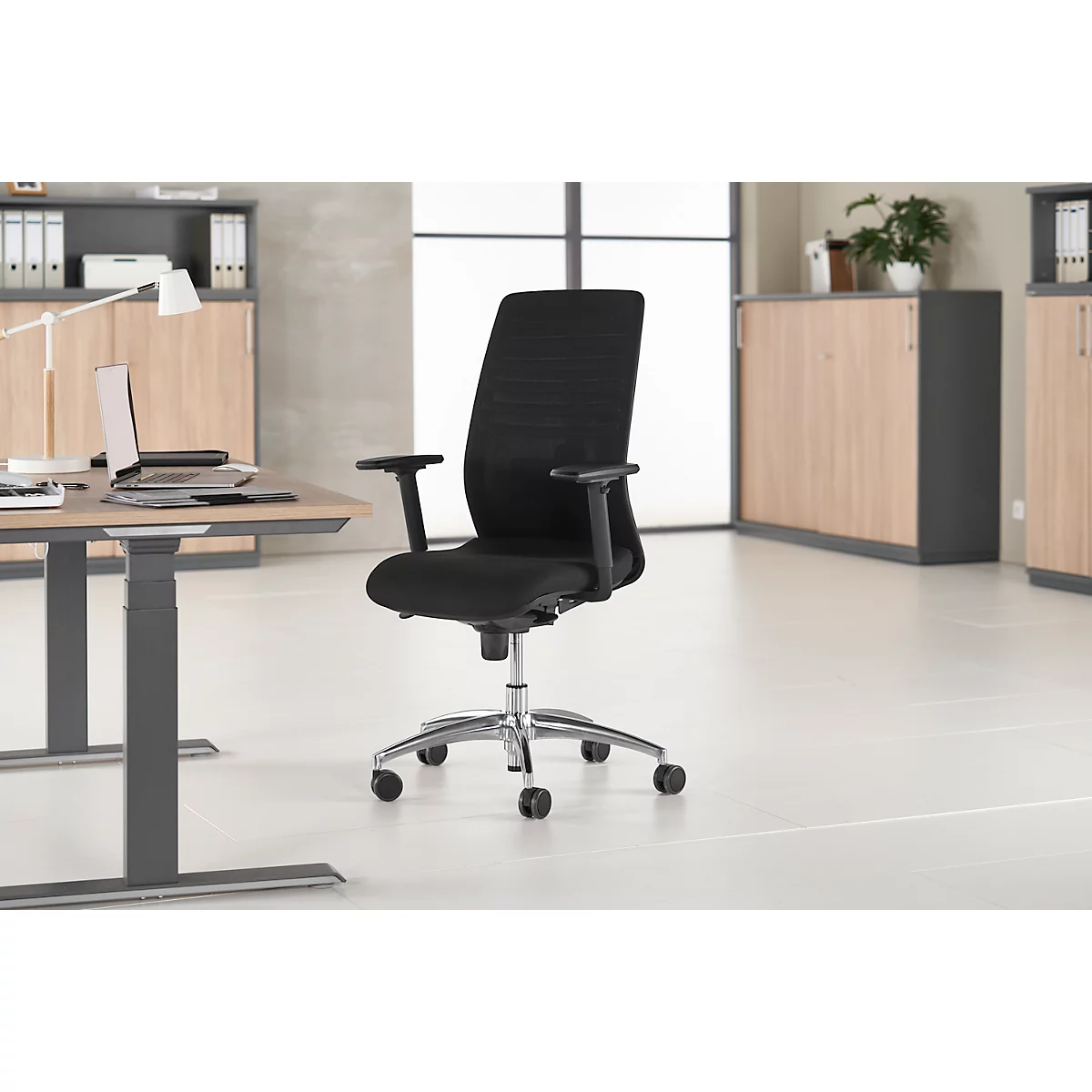 Black office chair with armrests and wheels in front of a desk and wall unit.