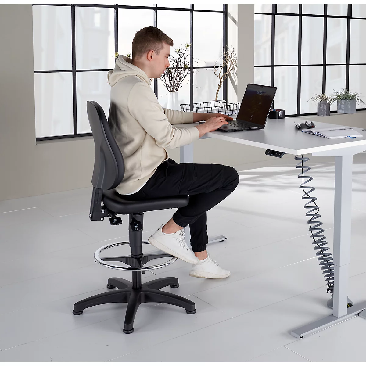 Un homme est assis à un bureau réglable en hauteur, travaillant sur un ordinateur portable. Il est assis sur une chaise pivotante noire avec repose-pieds. Le bureau est lumineux.