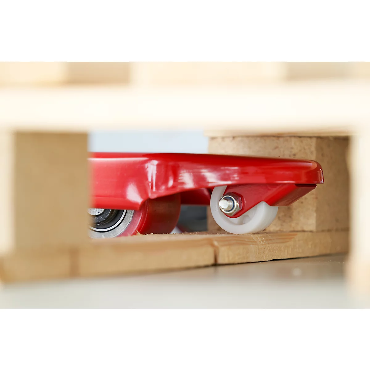 Detailaufnahme eines roten Transportrollers mit weißen Rollen, der teilweise in einem Holzrahmen sichtbar ist.