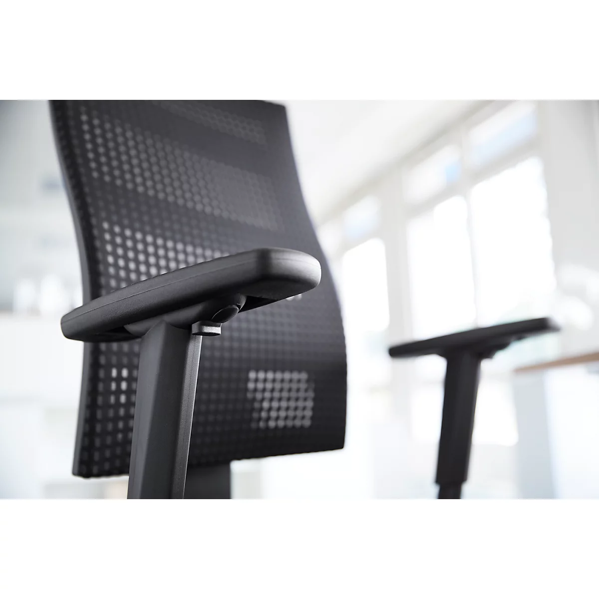 Black office chair with mesh back, armrest in focus. More chair parts and windows in the background.