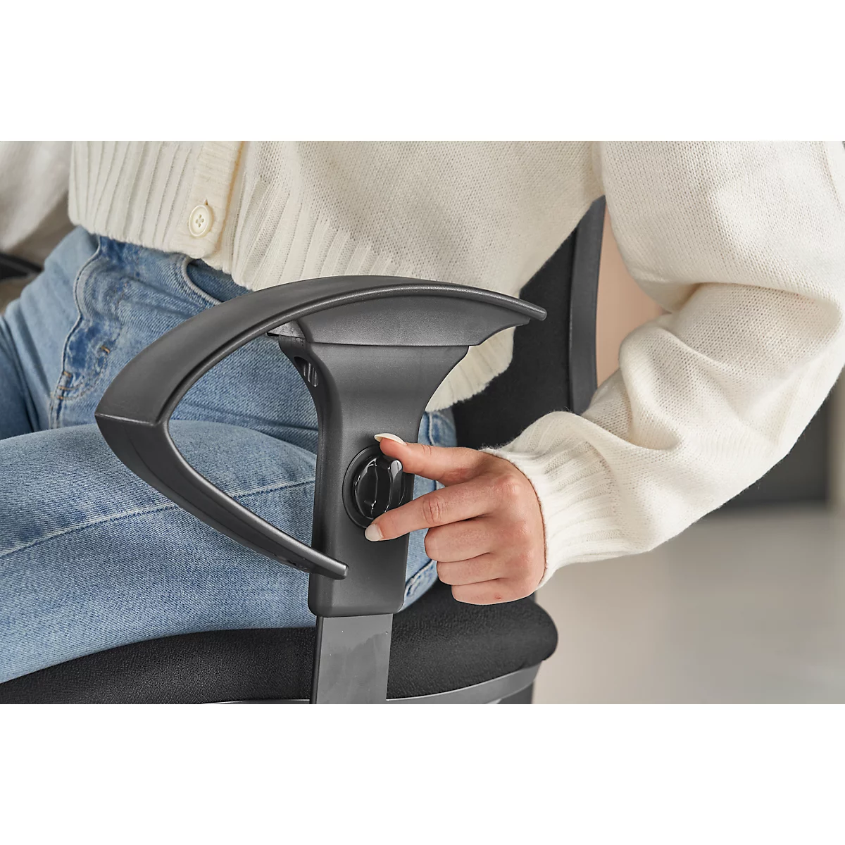 Close-up: Person adjusting the armrest of a black office chair. She is wearing a white sweater and jeans.