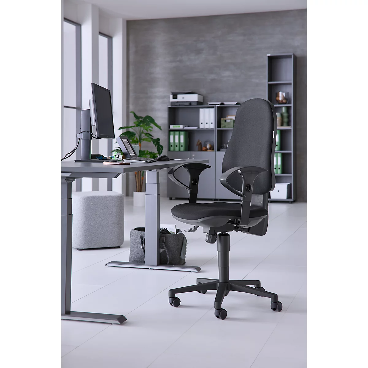 Office chair in front of a gray background. Desk, screen, plants, windows and shelves in the background. Light gray floor.