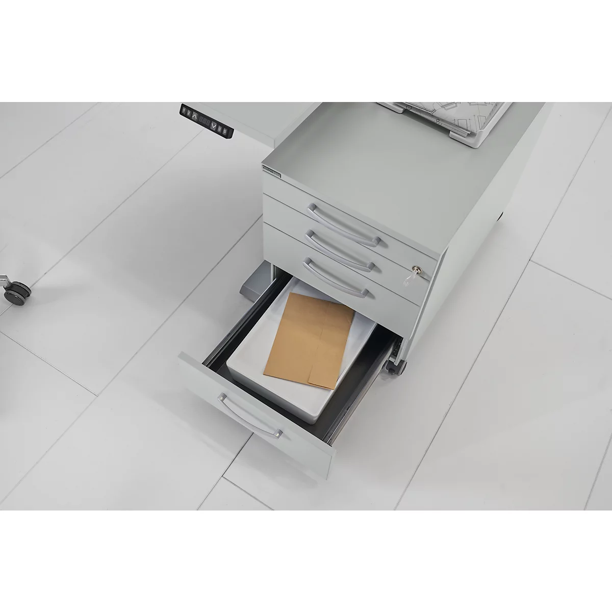 Grauer Rollcontainer mit drei Schubladen, eine davon geöffnet mit Papier. Auf hellem Boden, Draufsicht.